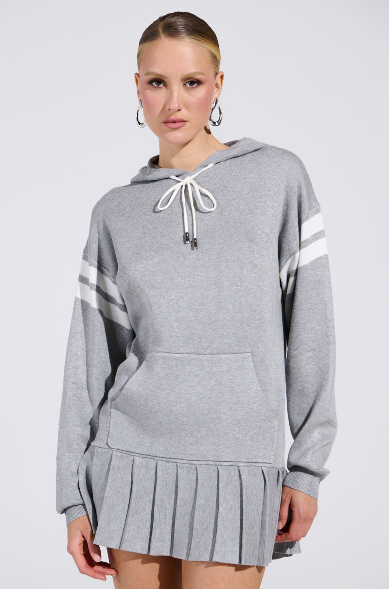 A person with tied-back hair in the BETTER OFF SWEATER JERSEY DRESS, a gray hoodie with white striped sleeves, pleated hem, mini silhouette, drawstring, and front pocket. They're wearing hoop earrings against a plain background.