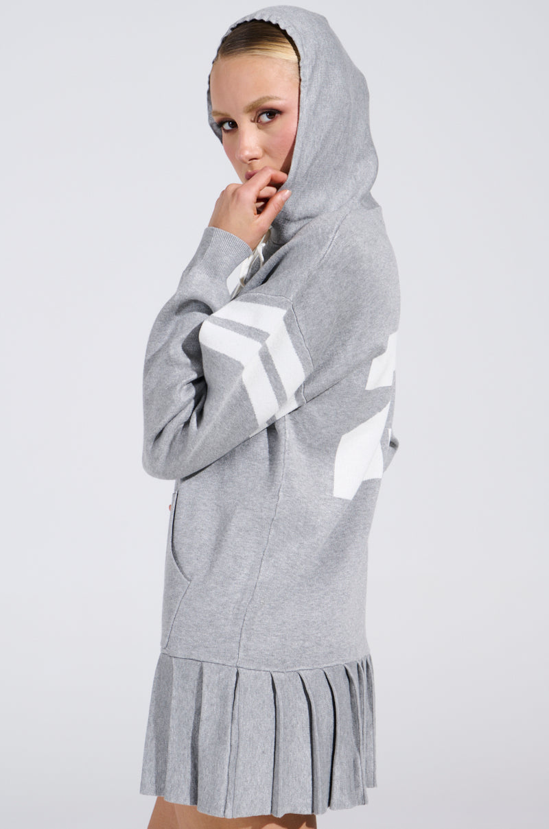 A person is wearing the BETTER OFF SWEATER JERSEY DRESS, a gray hooded dress with white geometric patterns on the sleeves and back. Featuring a knitted fabrication and pleated hem, they stand sideways with the hood up, gazing at the camera against a light gray background.