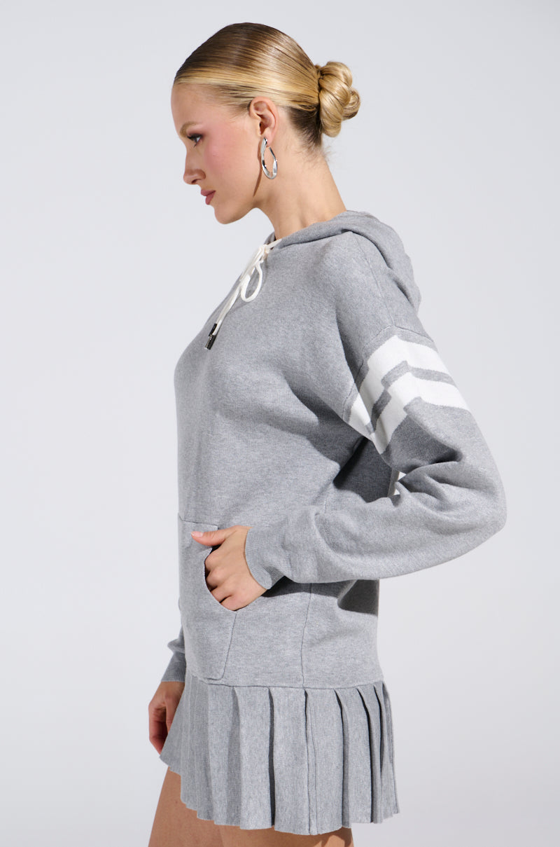 A blonde-haired woman in a bun wears the BETTER OFF SWEATER JERSEY DRESS, a gray hoodie style with white sleeve stripes and a pleated mini silhouette. Standing sideways with her hand in the pocket, she accents effortlessly with large hoop earrings.