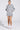 A person stands against a plain backdrop wearing the BETTER OFF SWEATER JERSEY DRESS in light gray, featuring white striped sleeves and a seamless pleated skirt. Their outfit is complemented by white lace socks, matching shoes, styled hair, and hoop earrings.