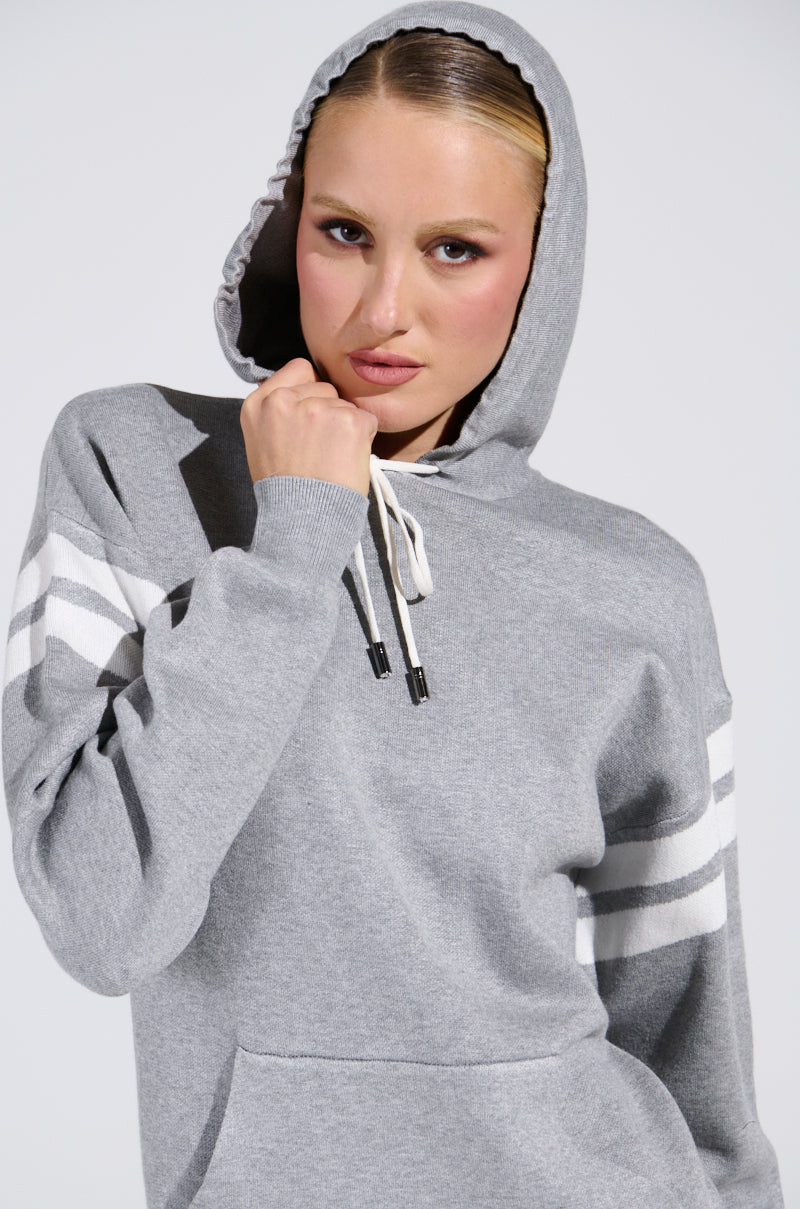 A person wearing the BETTER OFF SWEATER JERSEY DRESS, featuring white-striped sleeves and a hood up, poses with a serious expression. They grasp the knitted collar with one hand against a neutral background.