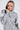 A person wearing the BETTER OFF SWEATER JERSEY DRESS, featuring white-striped sleeves and a hood up, poses with a serious expression. They grasp the knitted collar with one hand against a neutral background.