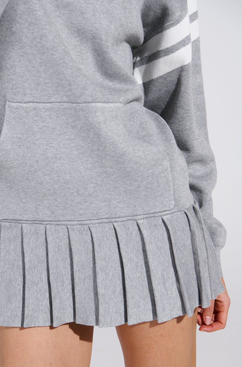 A person wears the BETTER OFF SWEATER JERSEY DRESS, a gray sweatshirt with white sleeve stripes and a pleated hem, against a soft gray background, focusing on the lower back and part of the arm.