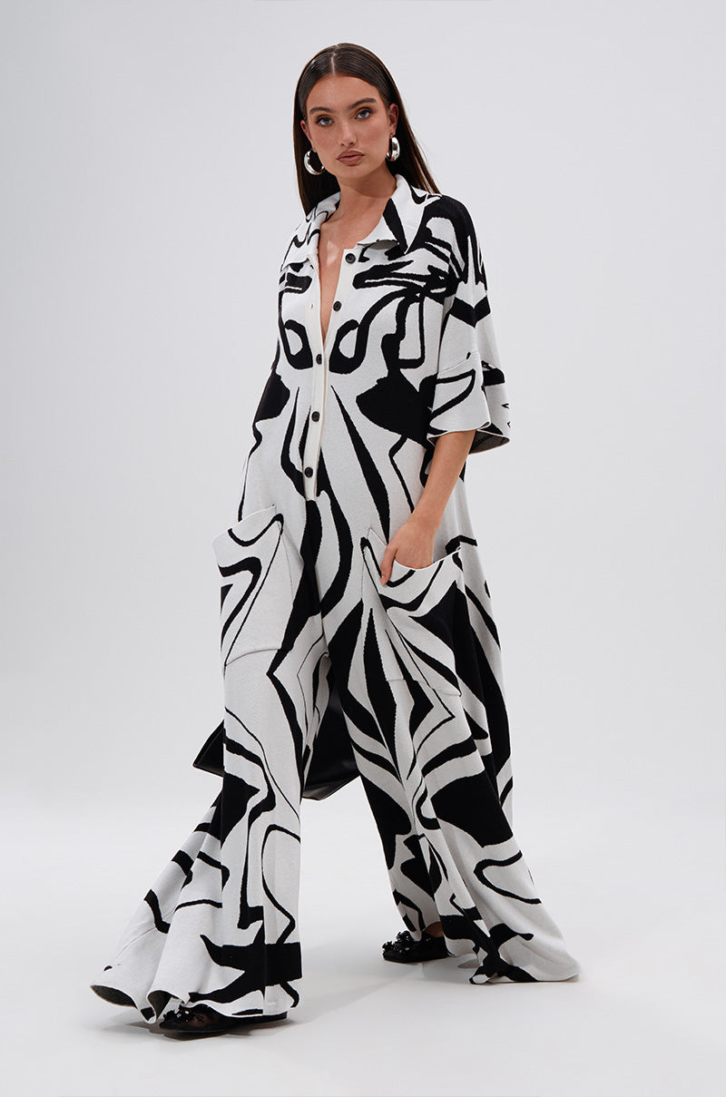 A woman poses in the HAPPY HOUR PRINTED KNIT JUMPSUIT by AKIRA, showcasing its black and white abstract print, wide legs, long sleeves, large pockets, and buttoned front against a plain light background.