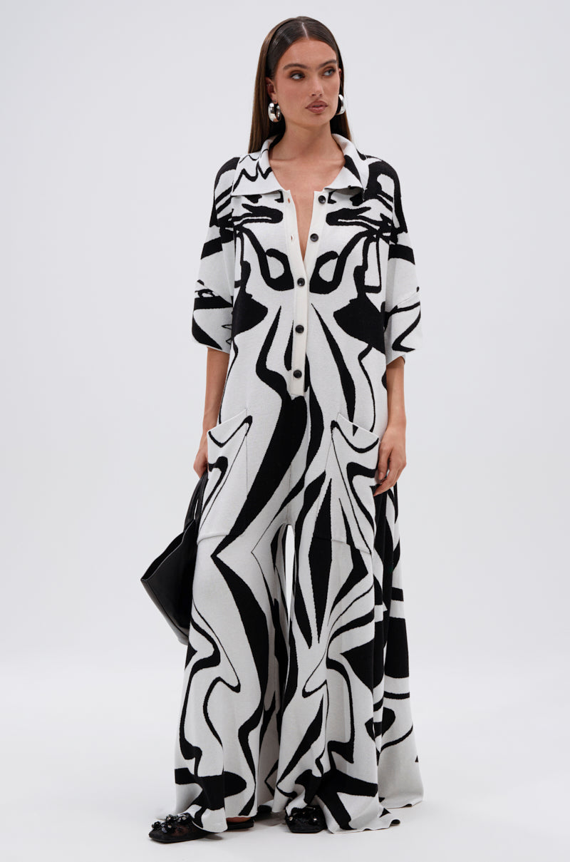 A woman wears the AKIRA Label HAPPY HOUR PRINTED KNIT JUMPSUIT, featuring a black and white abstract print and wide legs. She carries a black bag, sports long straight hair and statement earrings, standing against a plain white backdrop.