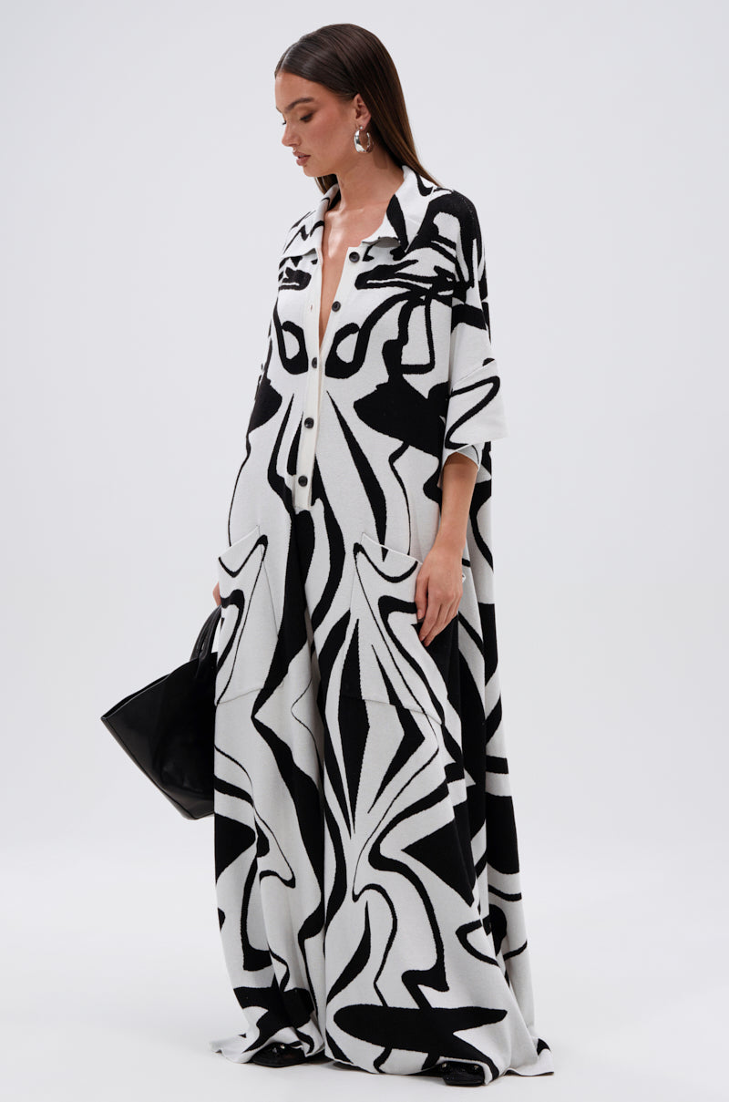 A woman wears the AKIRA Label HAPPY HOUR PRINTED KNIT JUMPSUIT with a bold abstract print, paired with a black handbag. Her straight brown hair and statement earrings complete her striking look.
