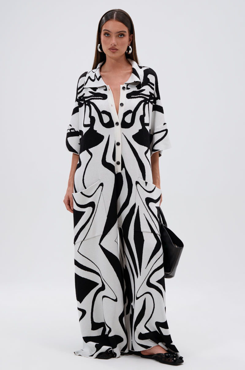 A woman in the HAPPY HOUR PRINTED KNIT JUMPSUIT, featuring a black-and-white abstract print, stands against a white background. She holds a black handbag and wears black shoes, her hair straight and tucked behind her ears.