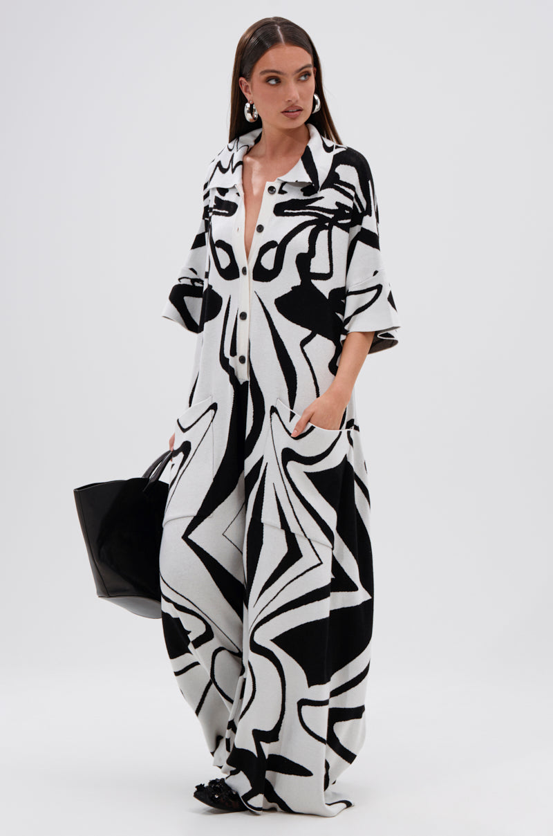 A woman wearing the AKIRA Label HAPPY HOUR PRINTED KNIT JUMPSUIT with a black and white abstract print and pockets stands against a white background, holding a large black bag and wearing statement earrings.