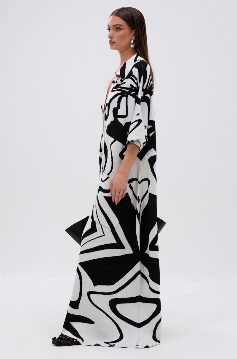 A woman stands in profile wearing the HAPPY HOUR PRINTED KNIT JUMPSUIT, featuring a bold black-and-white abstract print. She holds a black clutch, wears statement earrings, and poses with straight hair against a plain white background.