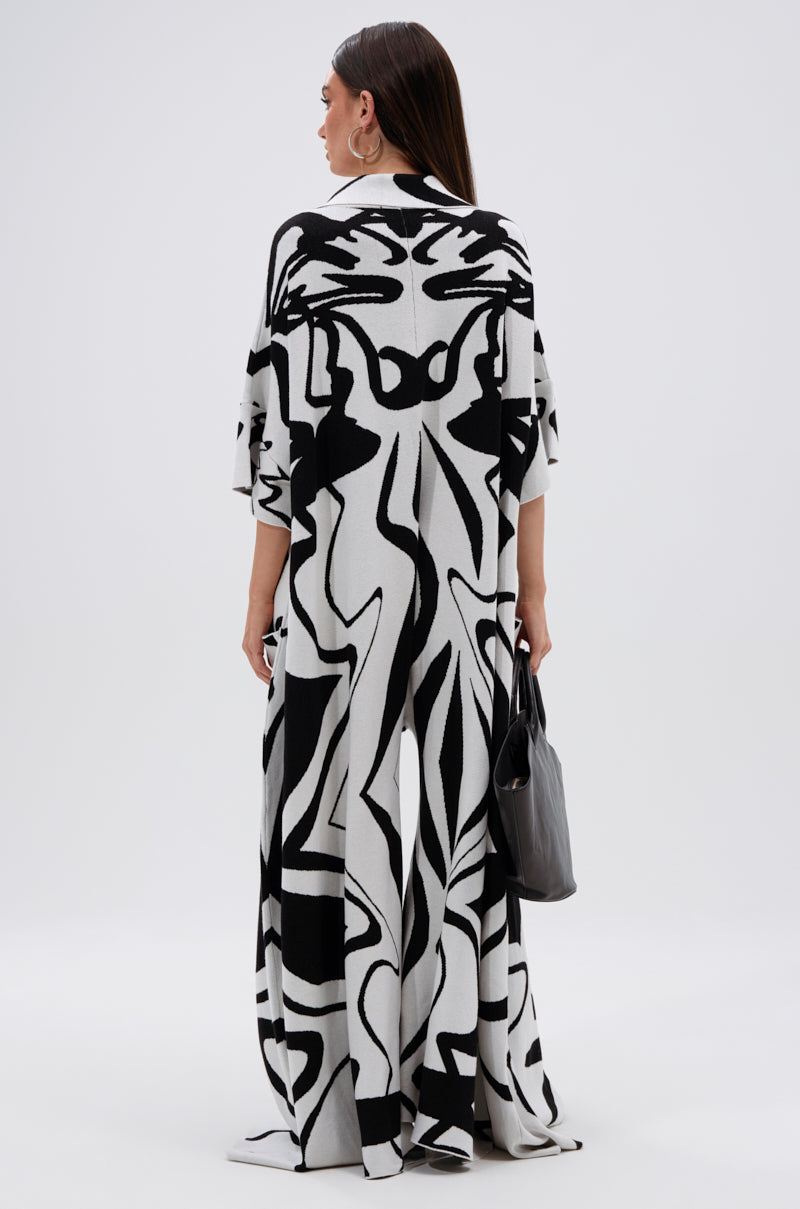 A woman faces away in the HAPPY HOUR PRINTED KNIT JUMPSUIT, featuring a black and white abstract print. She holds a gray handbag, with long straight hair and large hoop earrings, set against a plain light background.