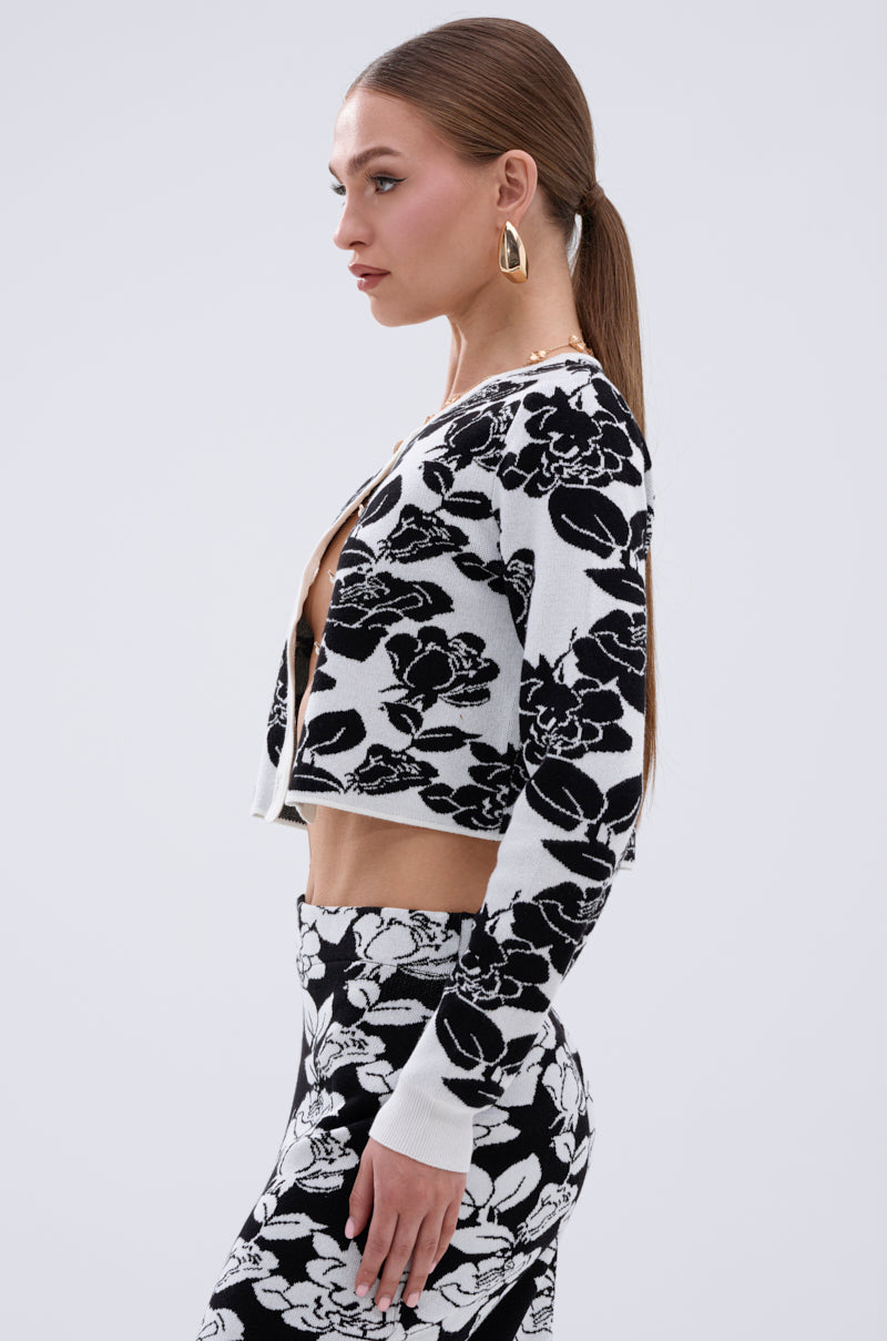 A woman with straight brown hair in a low ponytail wears gold geometric earrings and the BLACK ROSE PETAL CARDIGAN with pearl buttons, plus coordinating black-and-white floral pants, standing in profile against a plain light background.