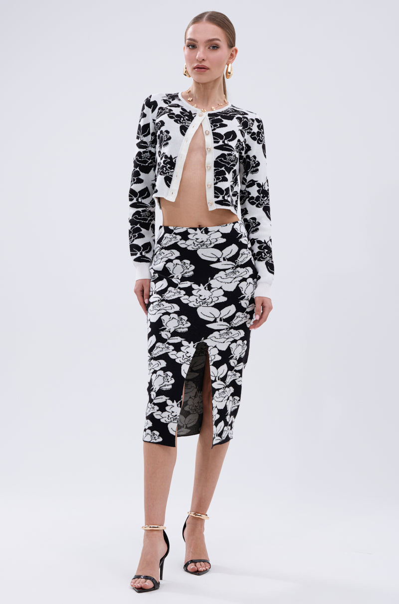 A woman wears the BLACK ROSE PETAL CARDIGAN with jacquard rose pattern and pearl buttons, paired with a matching skirt, black ankle-strap heels, and gold hoop earrings, standing against a plain white background.