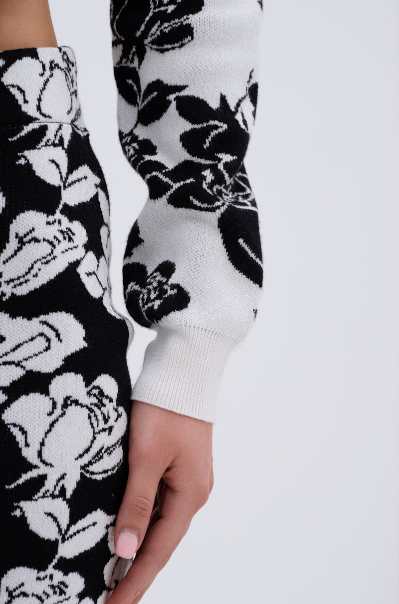A close-up shows a person's arm and hand wearing the BLACK ROSE PETAL CARDIGAN, featuring a jacquard rose pattern, with their hand resting at their side against a plain light background.