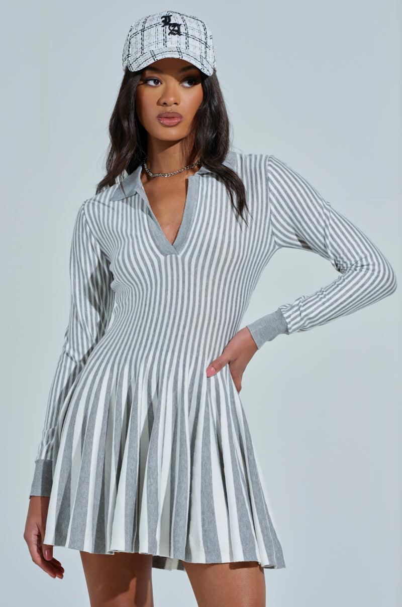 Wearing the ALINA pleated knit fit and flare mini dress in grey multi, a person poses confidently. They sport long dark hair, a white and black checkered cap, and a necklace against a soft gray background.
