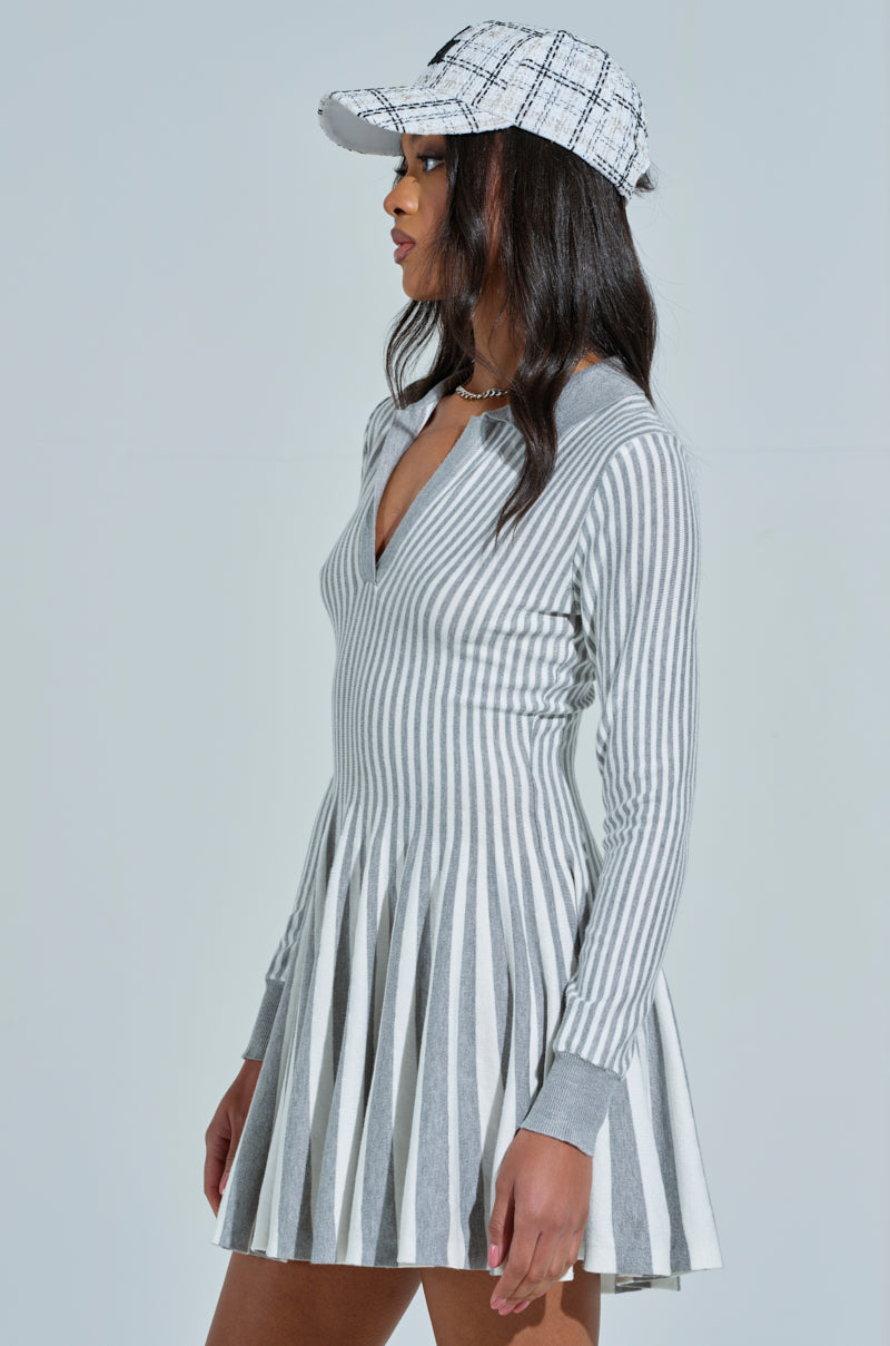 A woman poses in the ALINA Pleated Knit Fit and Flare Mini Dress with long sleeves in Grey Multi. Her plaid cap complements her side-turned head, with hair cascading below against a light background.