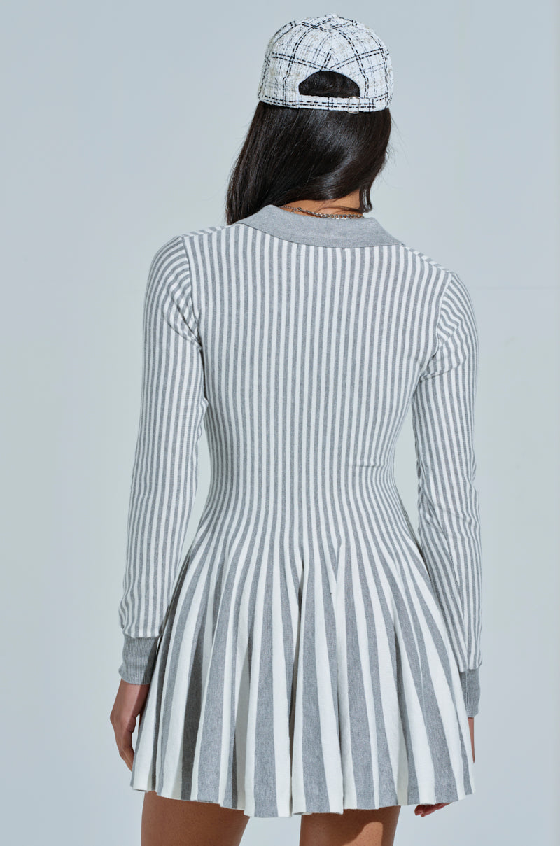 The elegance of the fabric drape is highlighted as a person wearing the ALINA Pleated Knit Fit and Flare Mini Dress with Long Sleeves in Grey Multi, along with a patterned cap, is shown from behind against a plain background.