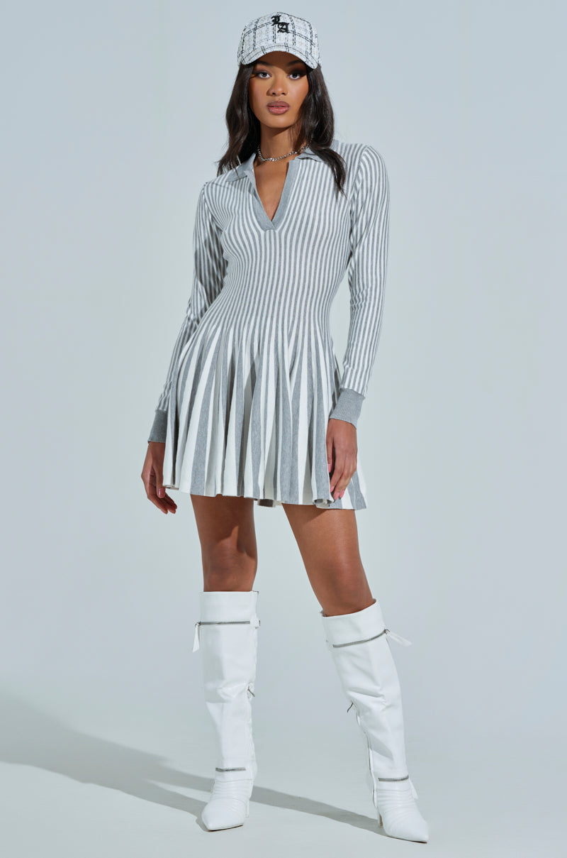Wearing the ALINA Pleated Knit Fit and Flare Mini Dress with long sleeves in grey multi, a person stands confidently against a simple background, paired with a plaid cap and knee-high white boots.