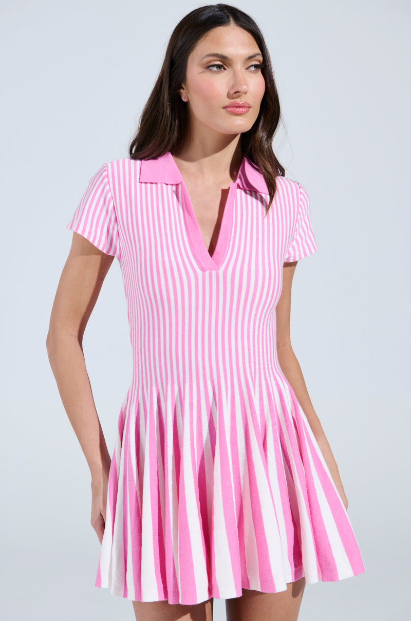 A woman in the ALINA Pleated Knit Fit and Flare Mini Dress in Pink Multi, with a collar and pleated skirt, stands against a plain backdrop. Her long dark hair cascades as she gazes slightly to the side.