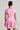 A person with dark hair styled in a sleek bun is shown from the back, wearing the ALINA PLEATED KNIT FIT AND FLARE MINI DRESS, a short-sleeve pink mini dress featuring striped patterning and a flared skirt. The person is holding a small, white polka-dotted handbag.