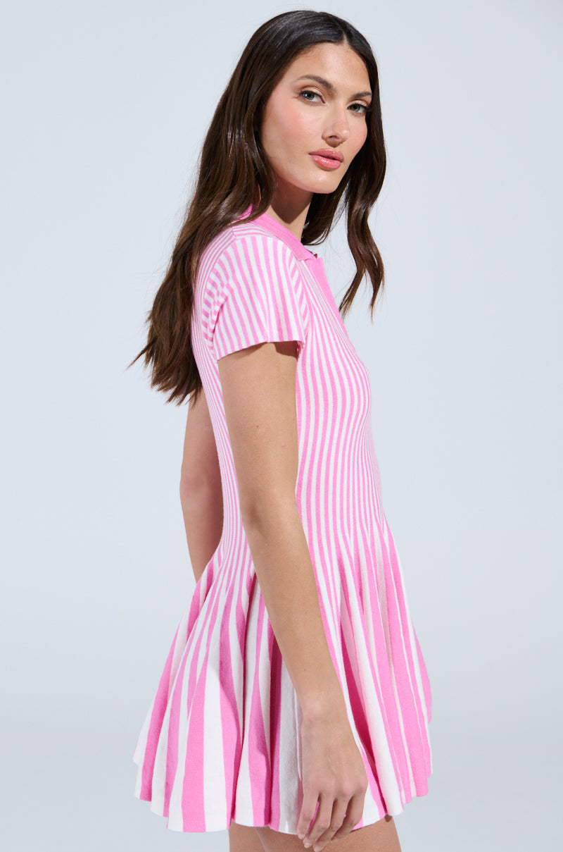 A person with long brown hair wears the ALINA Pleated Knit Fit and Flare Mini Dress in Pink Multi, featuring vertically striped short sleeves. The dress flares elegantly as they glance over their shoulder against a plain light background.