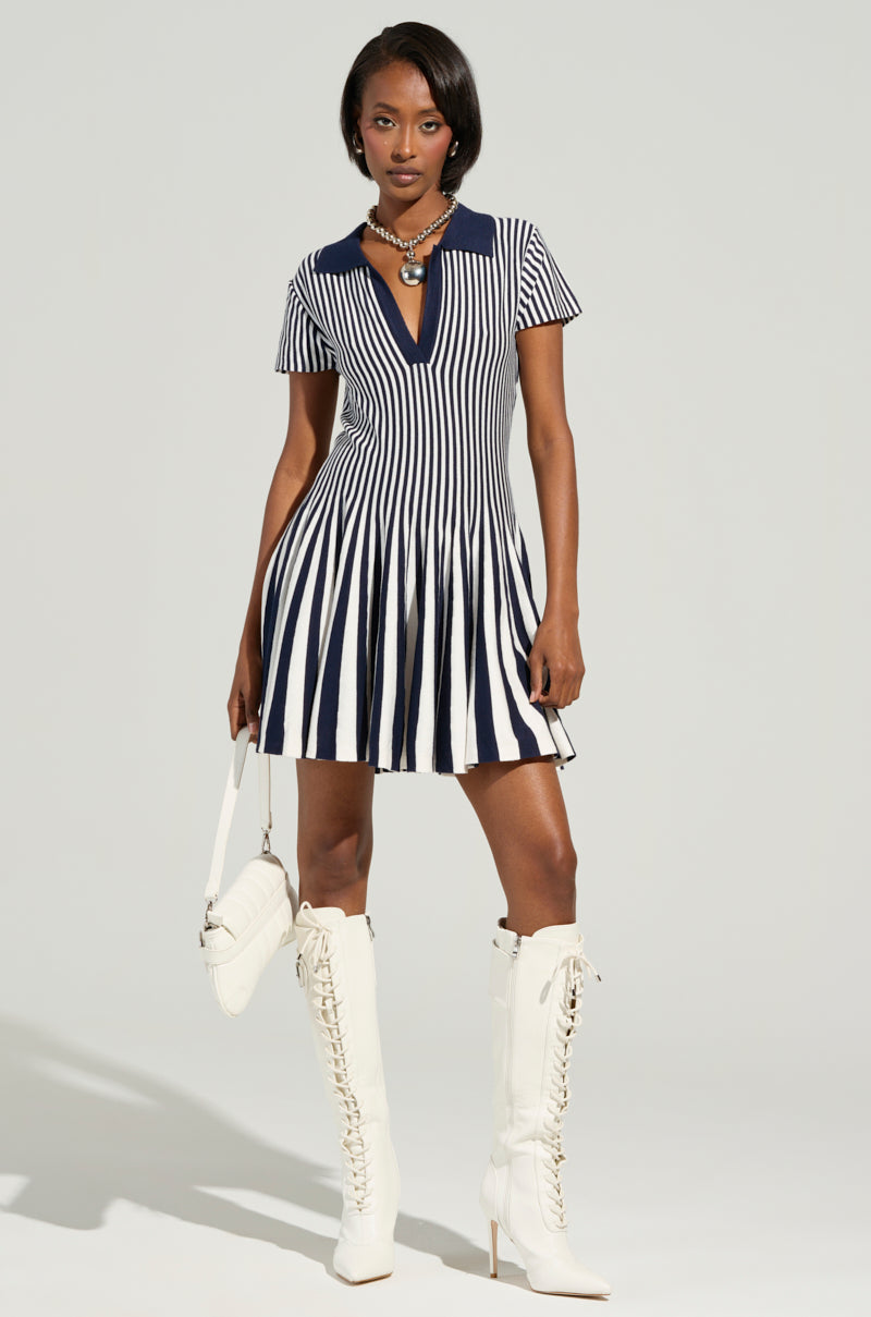 Wearing the Alina Pleated Knit Fit and Flare Mini Dress in Navy, a person stands confidently with knee-high white lace-up boots. They complement their look with a white purse and an elegant necklace, all set against a neutral background.