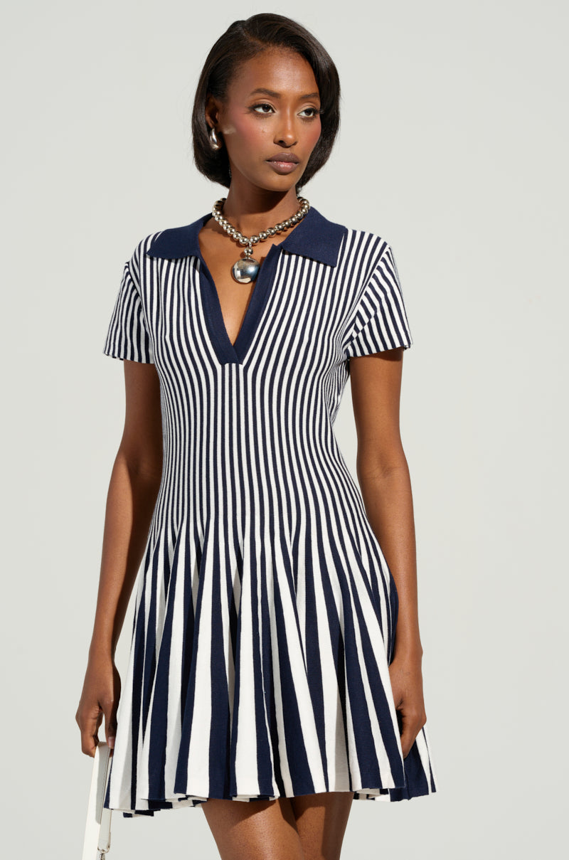 A woman wearing the Alina Pleated Knit Fit and Flare Mini Dress in Navy stands against a plain background. The bold, color-blocked striped design of the dress enhances her large necklace with a spherical pendant. Holding a white item, she exudes a poised and confident demeanor.