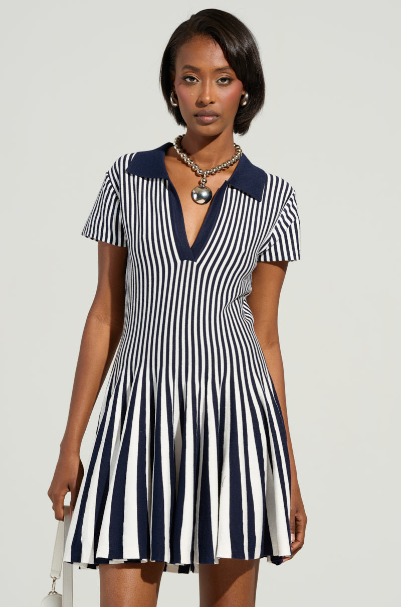 A person wears the ALINA Pleated Knit Fit and Flare Mini Dress in navy, featuring a collar and short sleeves, set against a plain background. Their shoulder-length hair complements the look, while a chunky necklace adds flair, enhancing the bold colorblock striped pattern of the outfit.