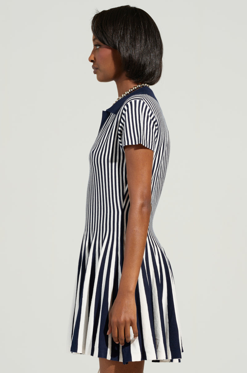 The individual is turned to the side, showcasing the ALINA Pleated Knit Fit and Flare Mini Dress in Navy, featuring a striking colorblock striped pattern for a vibrant effect. The simple backdrop draws attention to the contemporary and fashionable ensemble.