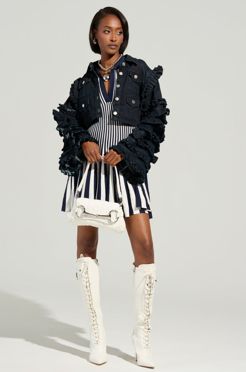 Wearing a ruffled denim jacket over the ALINA Pleated Knit Fit and Flare Mini Dress in Navy, paired with knee-high white lace-up boots, they confidently hold a white handbag and accessorize with a pearl necklace, all set against a plain background.