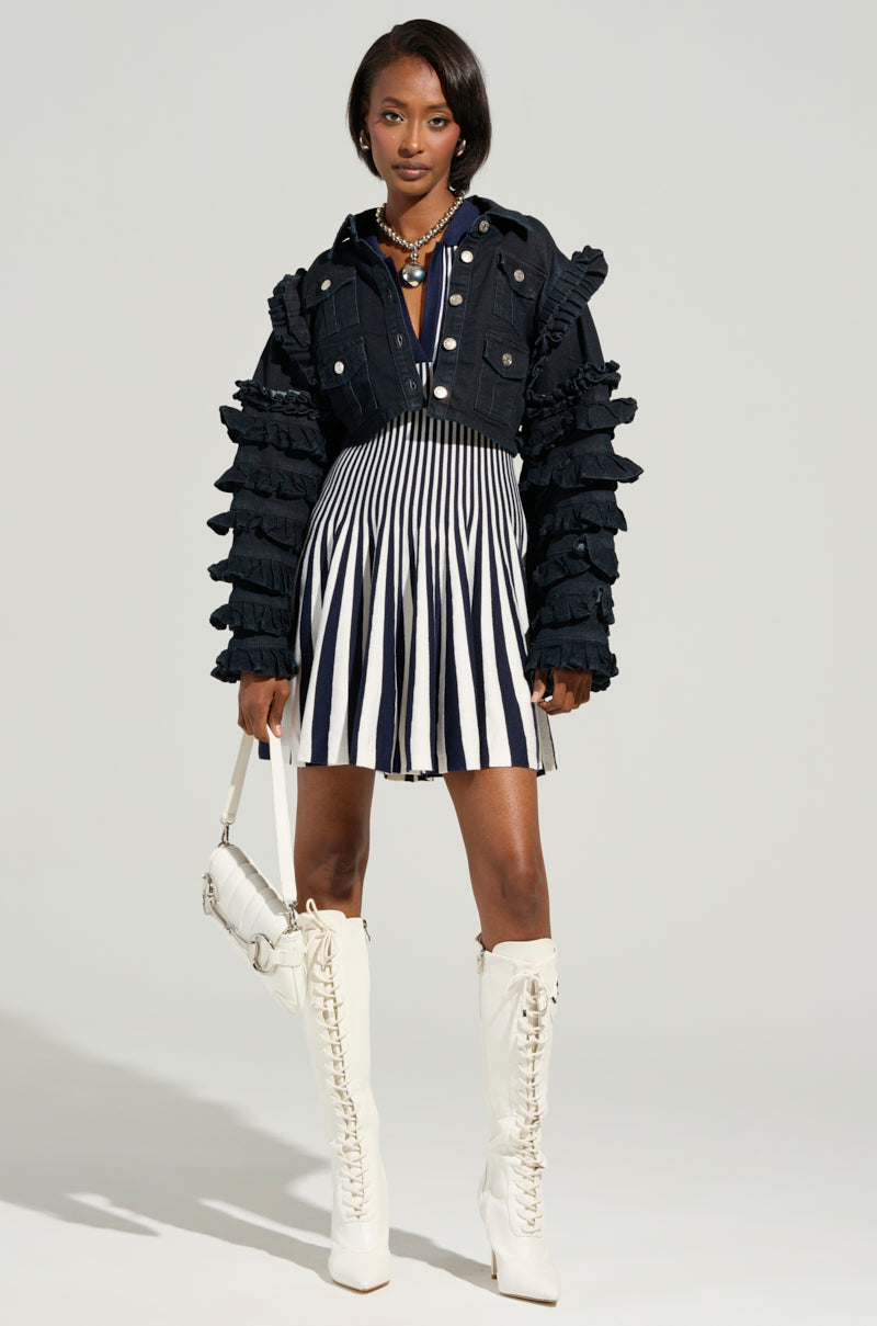 A woman stands confidently, donned in a navy Alina pleated knit fit and flare mini dress. She complements her stylish look with tall white lace-up boots, a white handbag, and layered necklaces. The neutral background enhances her chic ensemble.