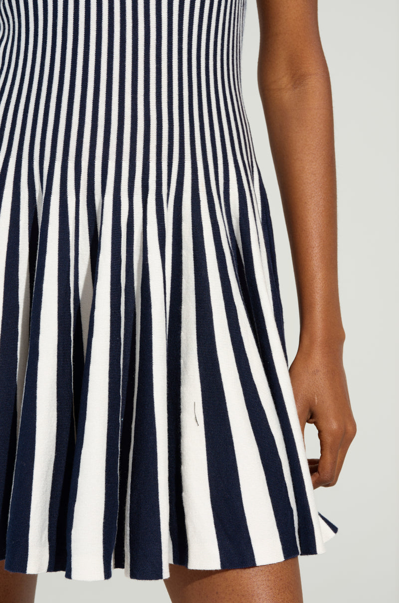 Close-up of a person wearing the Alina Pleated Knit Fit and Flare Mini Dress in Navy. The dress showcases a sleeveless design with a fitted bodice and pleated skirt in a vibrant colorblock striped pattern. One arm is visible, relaxed by their side, against a light, neutral background.