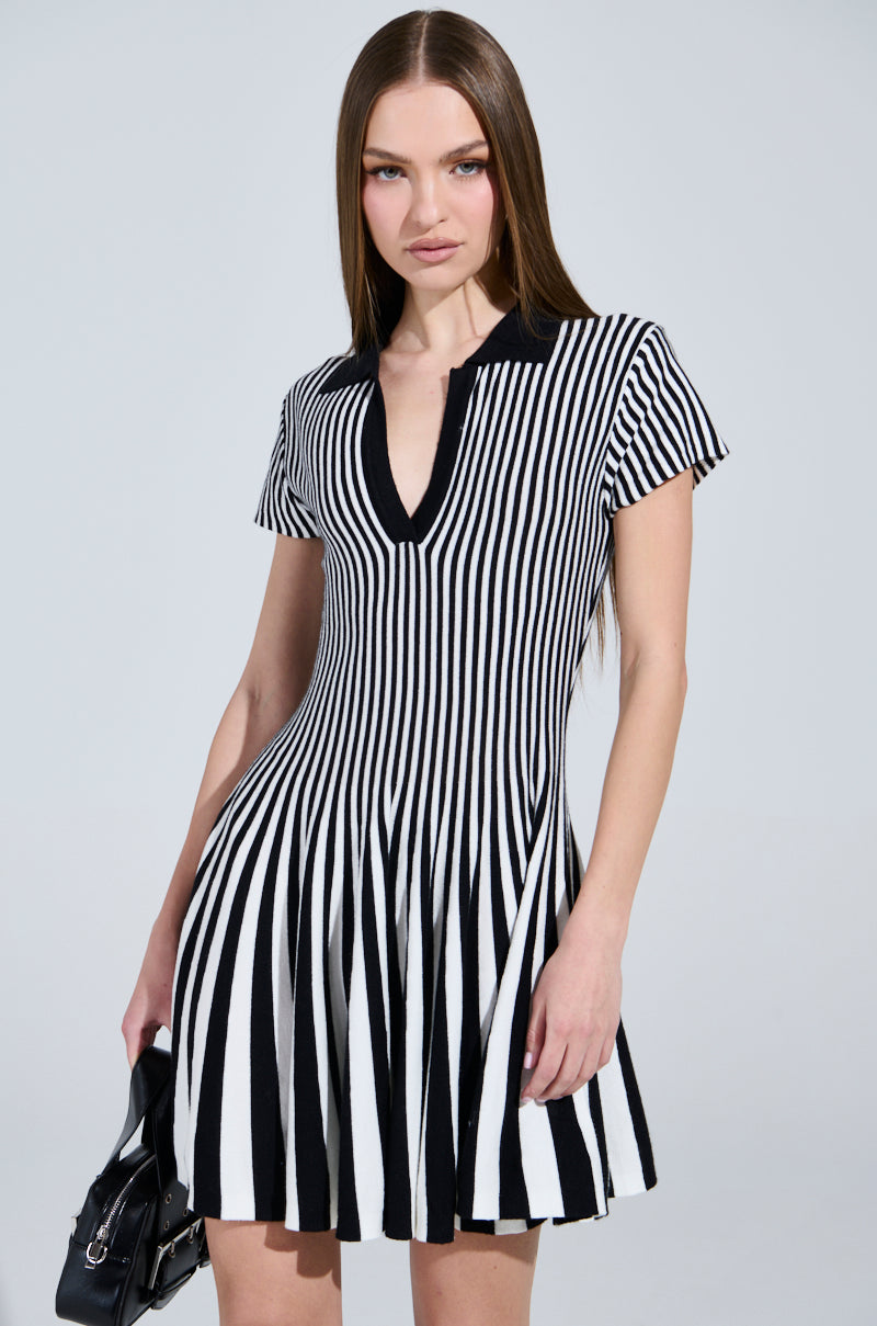 A person with long brown hair is wearing the ALINA Pleated Knit Fit and Flare Mini Dress in black and white. They hold a small black handbag, standing against a light gray background.