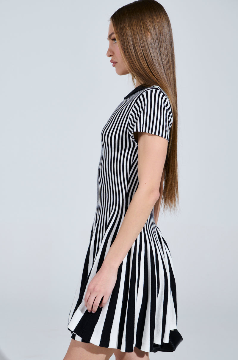 A person with long, straight hair wears the Alina Pleated Knit Fit and Flare Mini Dress in Black and White, featuring short sleeves. They stand in profile against a plain gray background.