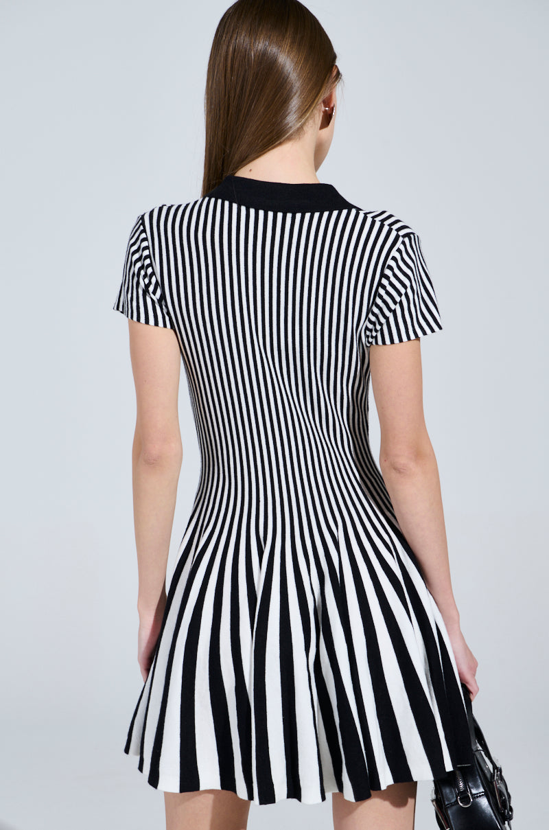 A person with long brown hair, shown from the back, is wearing the ALINA Pleated Knit Fit and Flare Mini Dress in black and white, featuring vertical stripes for a slimming effect and a flared skirt for added charm while holding a black handbag.