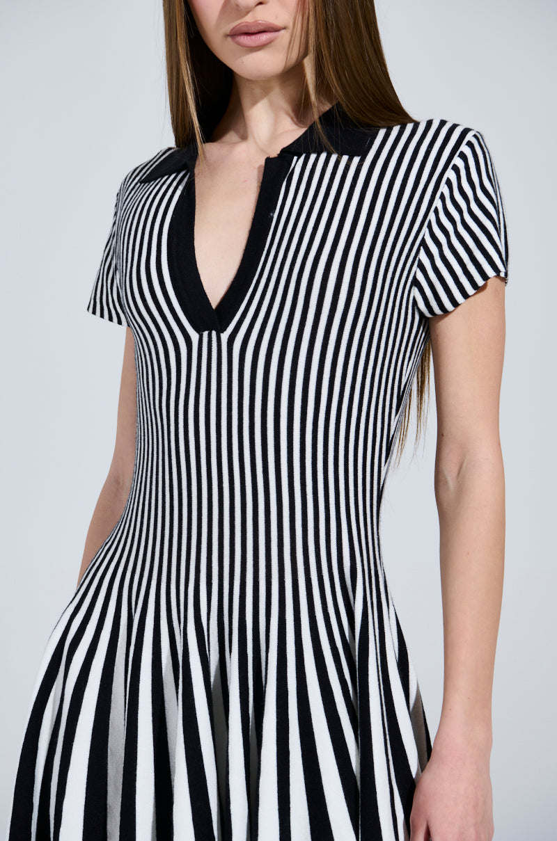 Someone wearing the ALINA PLEATED KNIT FIT AND FLARE MINI DRESS IN BLACK AND WHITE, featuring short sleeves and a V-neckline that creates an optical illusion. They have long, straight hair against a plain, light-colored background.