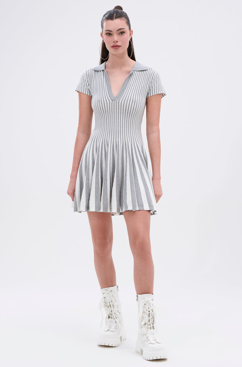 A woman poses against a white background in the ALINA PLEATED KNIT FIT AND FLARE MINI DRESS, featuring gray and white vertical stripes, a collared V-neck, skater silhouette, short sleeves, and paired with white lace-up boots.
