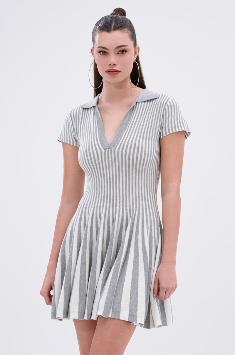 A woman with long dark hair in a half-up style wears the ALINA PLEATED KNIT FIT AND FLARE MINI DRESS—gray and white, vertically striped with a collar, V-neckline, and short sleeves. She accessorizes with large hoop earrings.