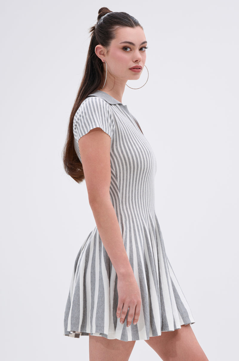 A woman with long brown hair in a half-up style wears the ALINA PLEATED KNIT FIT AND FLARE MINI DRESS, featuring gray and white vertical stripes and a flared skirt. She stands sideways, looking at the camera, with large hoop earrings.
