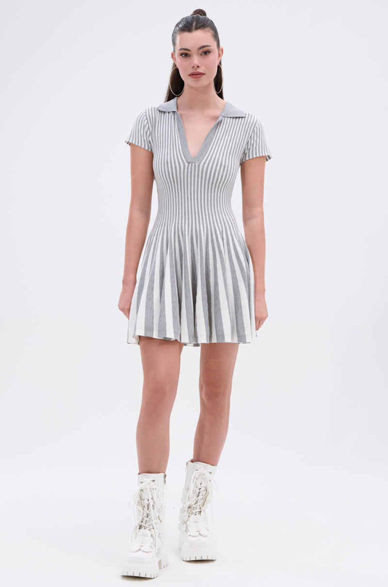 A woman with long dark hair tied back wears the ALINA PLEATED KNIT FIT AND FLARE MINI DRESS, featuring gray and white vertical stripes, short sleeves, a deep V-neck, and flared pleated skirt. She pairs it with white lace-up boots against a plain background.