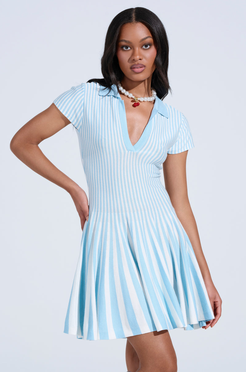 A woman models the ALINA PLEATED KNIT FIT AND FLARE MINI DRESS IN BLUE, a light blue and white vertically striped mini dress with short sleeves, V-neckline, and pleated skirt. She accessorizes with a necklace against a plain background.