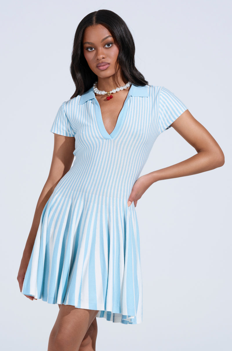 A woman wears the ALINA PLEATED KNIT FIT AND FLARE MINI DRESS IN BLUE, featuring light blue and white vertical stripes, short sleeves, a V-neck collar, and a flared skirt. She pairs it with a pearl necklace with a red pendant and has long, wavy hair.