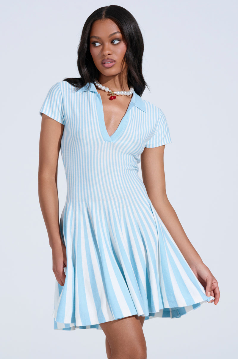 A woman wears the ALINA Pleated Knit Fit and Flare Mini Dress in blue, featuring short sleeves, a collared neckline, light blue and white vertical stripes, and a flared skirt. She accessorizes with a pearl necklace with a red pendant.