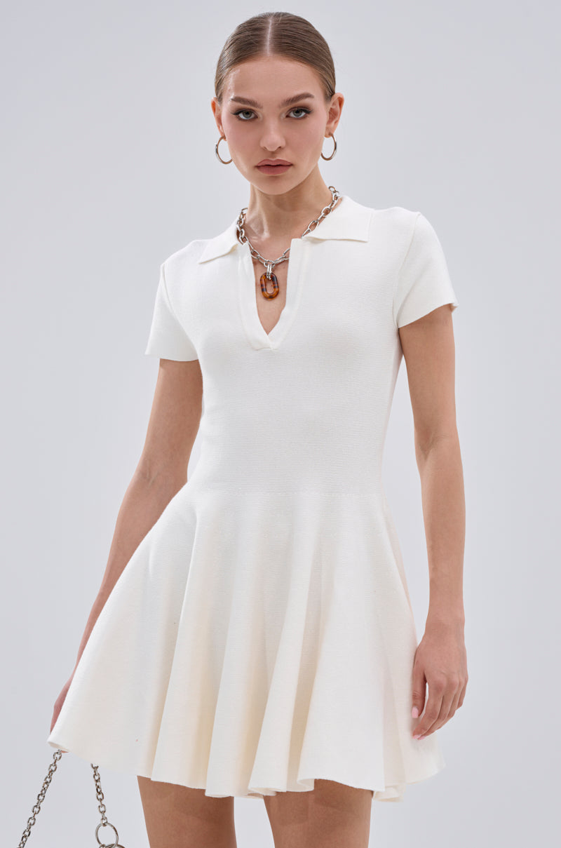 A woman wears the SLEEK AND CHIC KNIT MINI DRESS IN WHITE with short sleeves, a collar, and V-neckline. She pairs it with hoop earrings, a chunky chain necklace, and carries a stylish chain-strap bag. 0