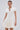 A woman wears the SLEEK AND CHIC KNIT MINI DRESS IN WHITE with short sleeves, a collar, and V-neckline. She pairs it with hoop earrings, a chunky chain necklace, and carries a stylish chain-strap bag.