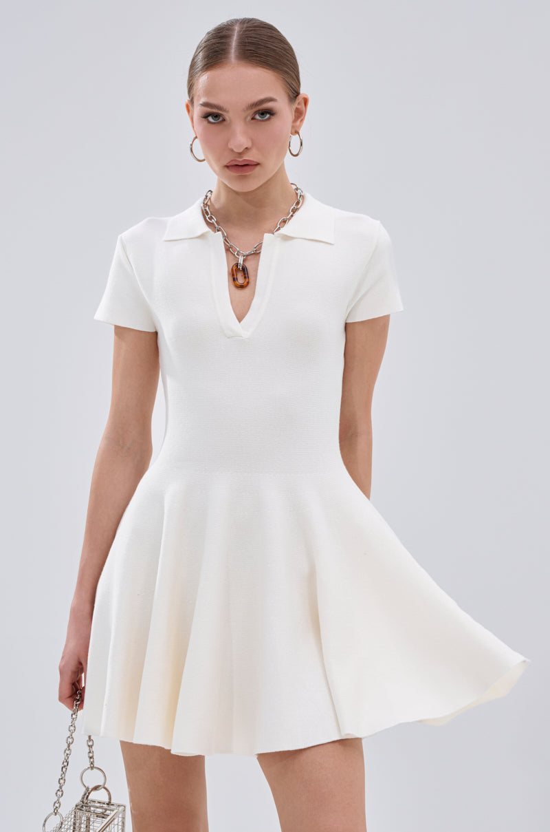 A woman wears the SLEEK AND CHIC KNIT MINI DRESS IN WHITE with short sleeves and a V-neck, accessorized with a chunky necklace and holding a small silver bag against a plain light background. 1