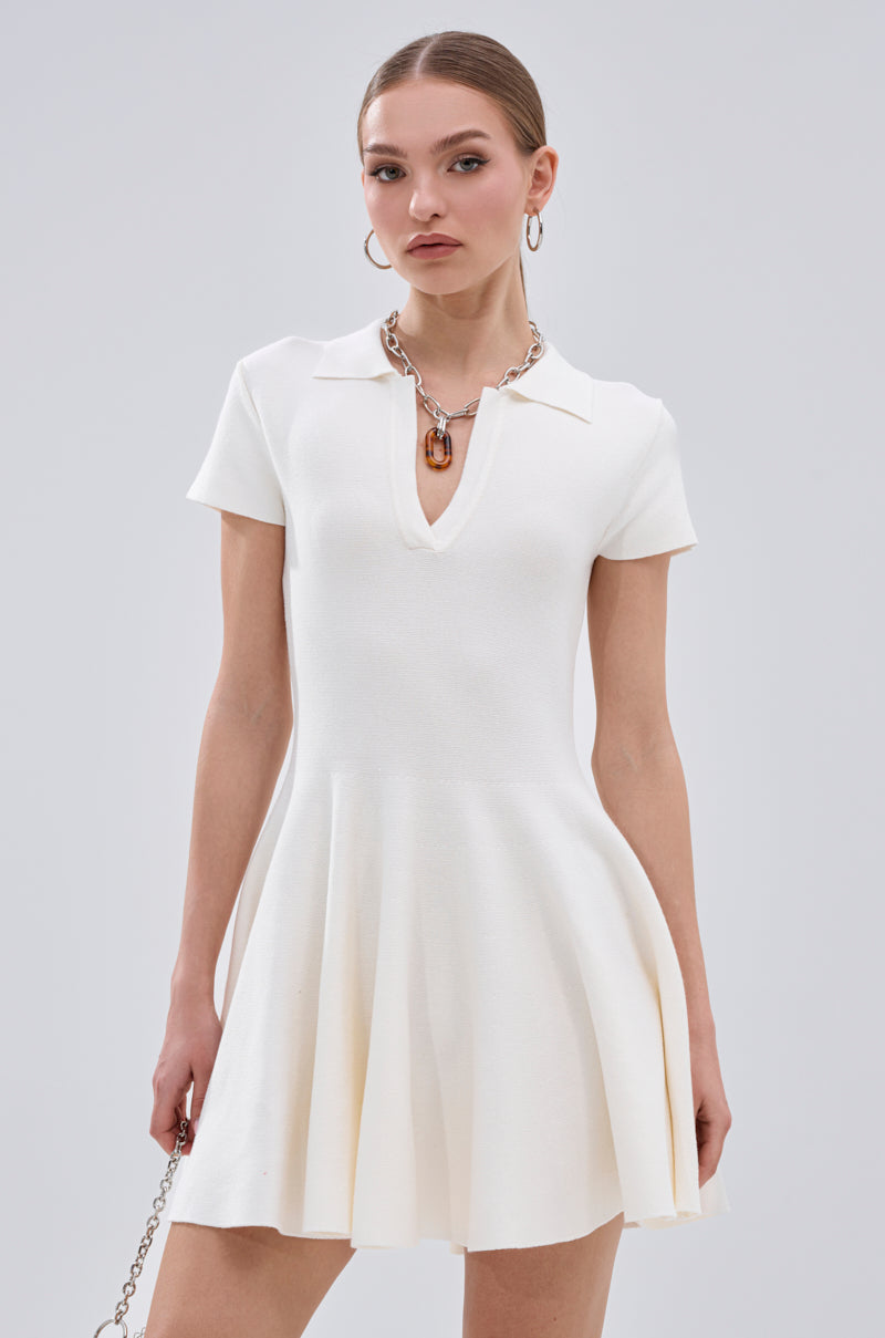 A woman wears the SLEEK AND CHIC KNIT MINI DRESS IN WHITE with short sleeves, a collared deep V-neckline, and flared skirt. She accessorizes with hoop earrings, a necklace, and a chain strap purse against a plain background. 2