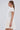 A woman with long, straight brown hair in a low ponytail wears the SLEEK AND CHIC KNIT MINI DRESS IN WHITE with short sleeves. She stands in profile against a plain light grey background, accessorized with hoop earrings and a chain necklace.