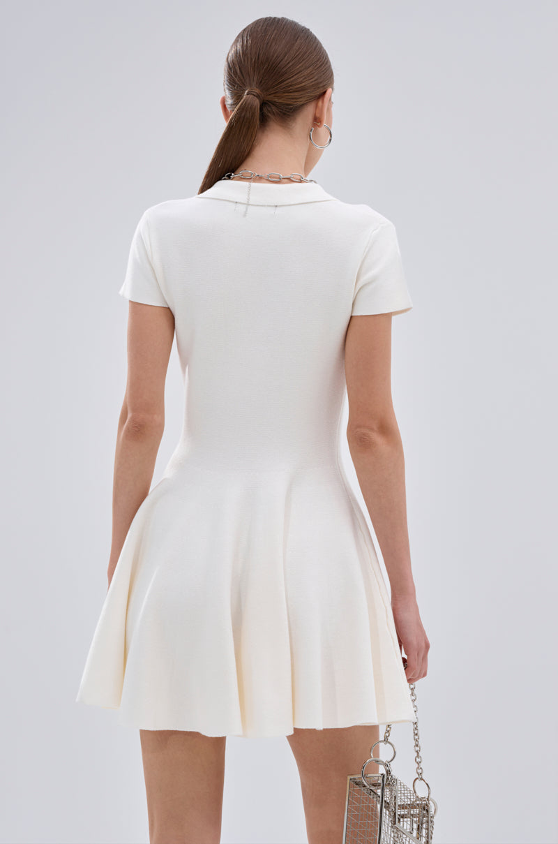 A woman with long brown hair tied back wears the SLEEK AND CHIC KNIT MINI DRESS IN WHITE. She stands with her back to the camera, holding a small metallic handbag in her left hand. 4