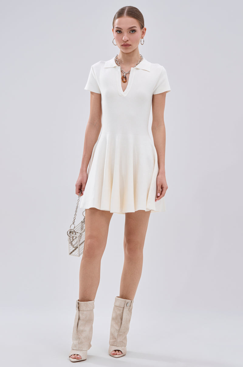 A woman stands against a light background wearing the SLEEK AND CHIC KNIT MINI DRESS IN WHITE. She pairs it with a small chain-strap purse, beige open-toe boots, and statement jewelry for a chic look. 5