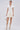 A woman stands against a light background wearing the SLEEK AND CHIC KNIT MINI DRESS IN WHITE. She pairs it with a small chain-strap purse, beige open-toe boots, and statement jewelry for a chic look.